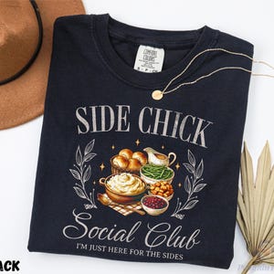 May include: Black crew neck t-shirt with the text "SIDE CHICK Social Club" and an illustration of Thanksgiving side dishes. The shirt also has the text "I'M JUST HERE FOR THE SIDES". A gold necklace and a brown hat are also in the image.