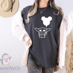 A woman wearing a black t-shirt with a white graphic of a cartoon character and a balloon, along with a white cardigan, standing in front of a wall with a hat and a penguin logo.
