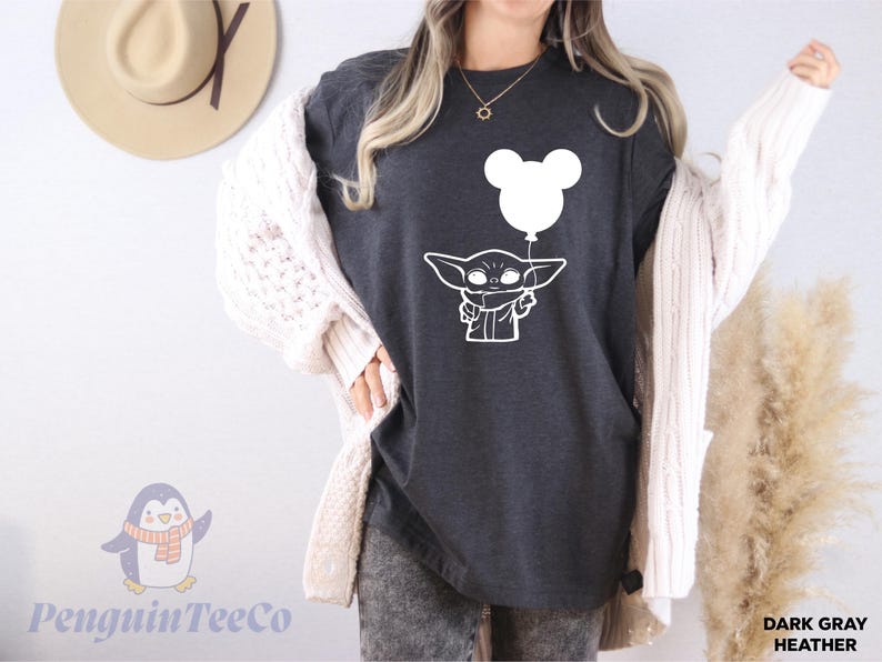 A woman wearing a black t-shirt with a white graphic of a cartoon character and a balloon, along with a white cardigan, standing in front of a wall with a hat and a penguin logo.
