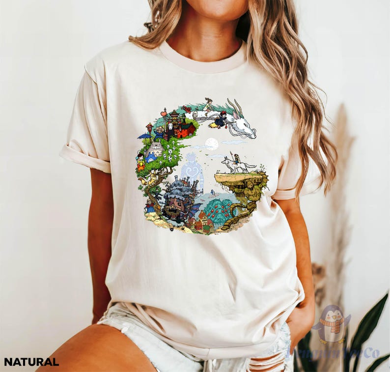 A woman wearing a white t-shirt with a colorful, illustrated design depicting a fantastical landscape with various elements such as buildings, animals, and a dragon.