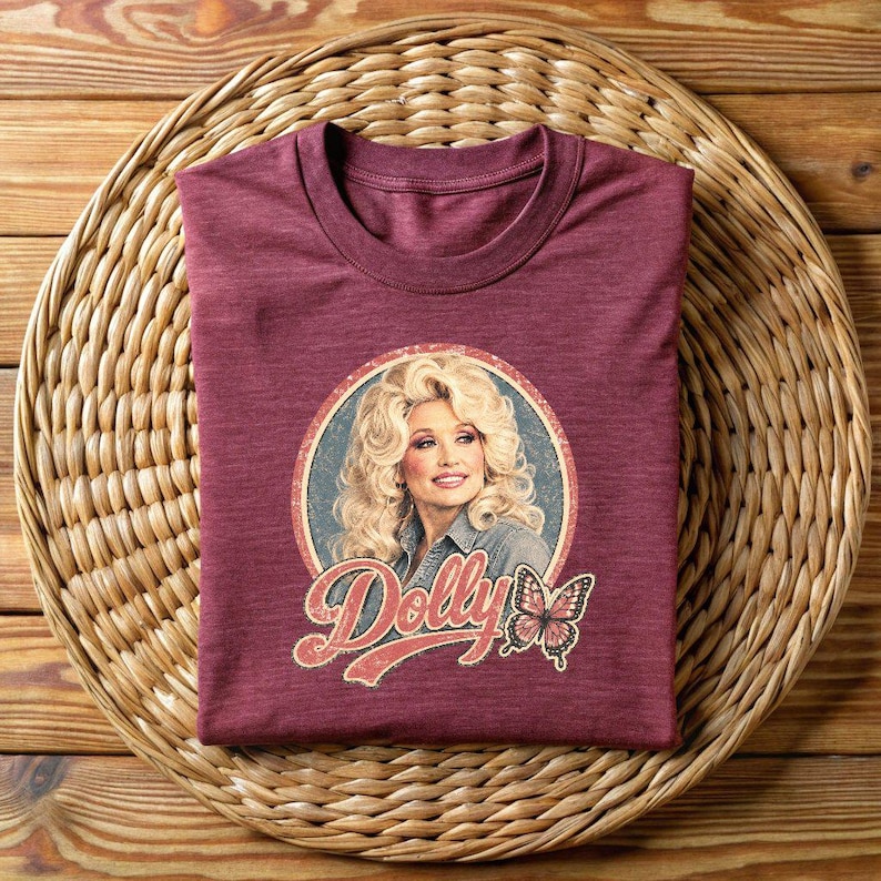 May include: A maroon crew neck t-shirt featuring a circular graphic of a blonde woman with voluminous hair wearing a denim shirt. Below the portrait, the name Dolly is printed in a stylized red font, accompanied by a small, detailed butterfly illustration.