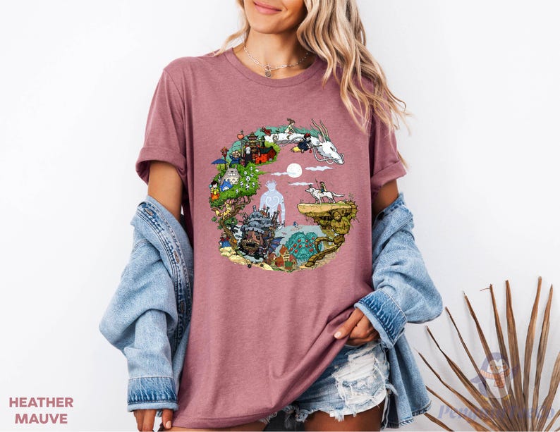 A woman wearing a pink t-shirt with a colorful graphic design, including a castle, animals, and a moon. She is also wearing a denim jacket and denim shorts.