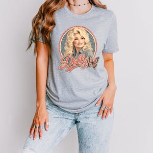 May include: A light gray short-sleeved t-shirt featuring a circular graphic print of a blonde person with the text Dolly in a retro font and a butterfly illustration. The design is centered on the chest of the shirt, which is worn with light blue jeans.