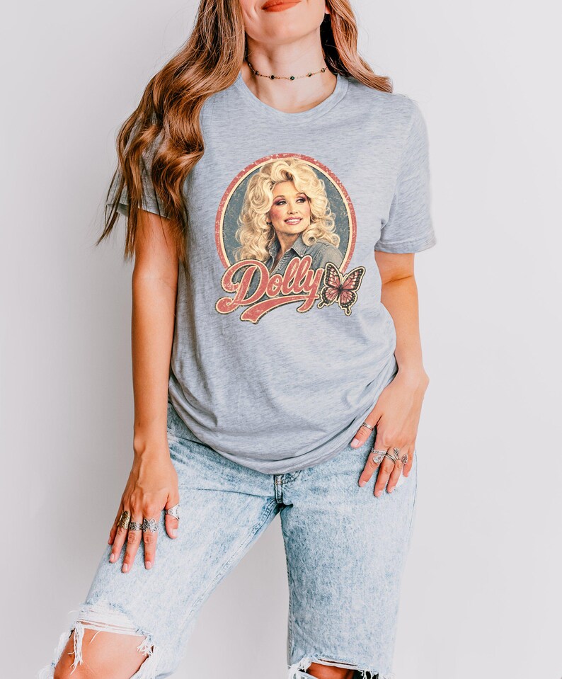 May include: A light gray short-sleeved t-shirt featuring a circular graphic print of a blonde person with the text Dolly in a retro font and a butterfly illustration. The design is centered on the chest of the shirt, which is worn with light blue jeans.