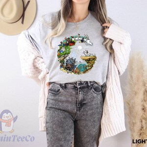 A woman wearing a gray t-shirt with a colorful graphic design, gray jeans, and a white cardigan stands in front of a wall with a hat and a potted plant.