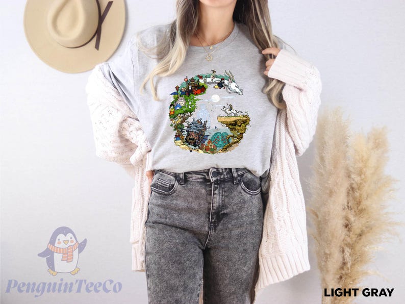 A woman wearing a gray t-shirt with a colorful graphic design, gray jeans, and a white cardigan stands in front of a wall with a hat and a potted plant.