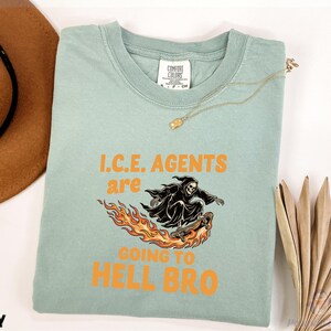 May include: A light green Comfort Colors t-shirt with the text "I.C.E. AGENTS ARE GOING TO HELL BRO" in orange. The graphic shows a black Grim Reaper skateboarding on flames. A gold necklace is draped on the shirt.