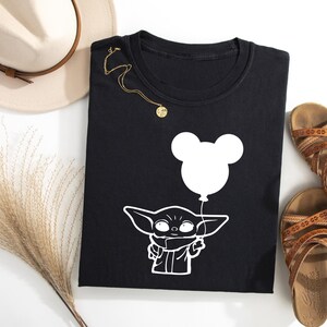 a black t-shirt with a white graphic of a baby alien character holding a balloon, along with a beige hat, brown sandals, and a decorative plant.