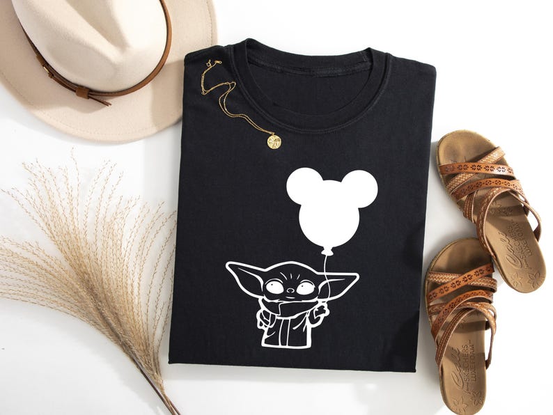 a black t-shirt with a white graphic of a baby alien character holding a balloon, along with a beige hat, brown sandals, and a decorative plant.