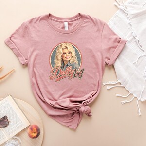 May include: A mauve short-sleeve t-shirt featuring a circular graphic print of a blonde woman with the text Dolly in a stylized red font, accompanied by a butterfly illustration. The shirt has a crew neckline and a knotted hem detail at the side.