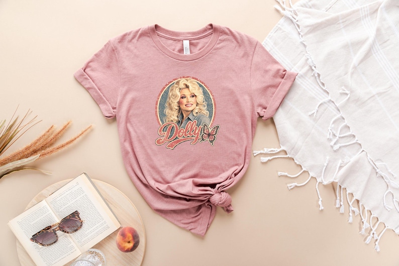 May include: A mauve short-sleeve t-shirt featuring a circular graphic print of a blonde woman with the text Dolly in a stylized red font, accompanied by a butterfly illustration. The shirt has a crew neckline and a knotted hem detail at the side.