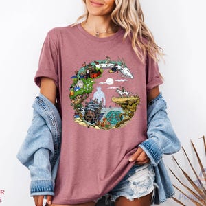 A woman wearing a pink t-shirt with a colorful graphic design, including a castle, animals, and a moon. She is also wearing a denim jacket and denim shorts.