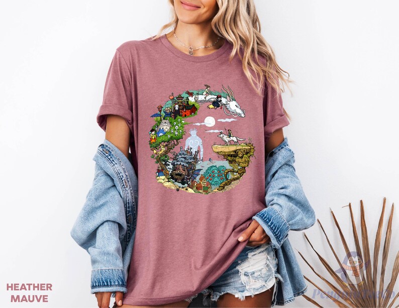 A woman wearing a pink t-shirt with a colorful graphic design, including a castle, animals, and a moon. She is also wearing a denim jacket and denim shorts.