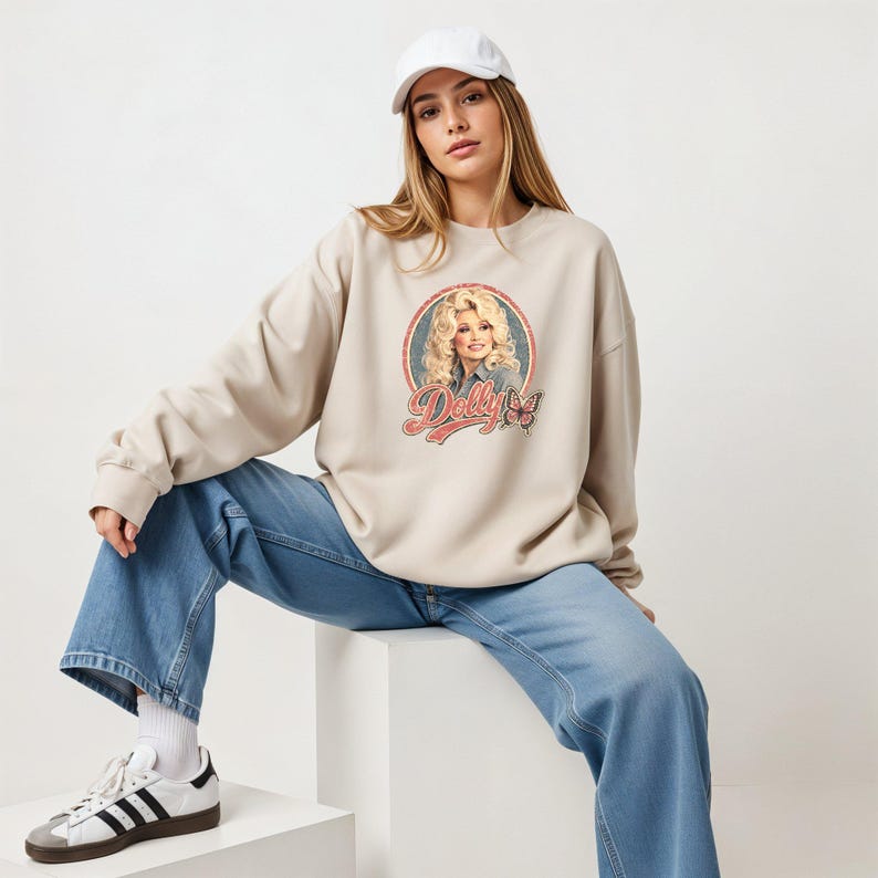 May include: A beige crewneck sweatshirt featuring a circular graphic print of Dolly Parton with blonde hair, a butterfly, and the text "Dolly" in a red script font. The sweatshirt is paired with blue denim jeans, a white baseball cap, and sneakers.