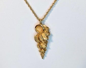 May include: A gold necklace with a seashell pendant. The pendant has a detailed, textured surface and a spiral design. The necklace is displayed on a white bust, highlighting the pendant's shape and colour.