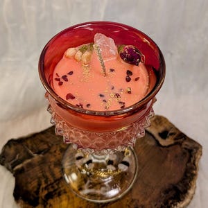 May include: A decorative candle in a red glass with a crystal and dried flowers. The candle is peach-colored and has gold flecks and small dark pieces. The glass has a decorative base and sits on a wooden slab.