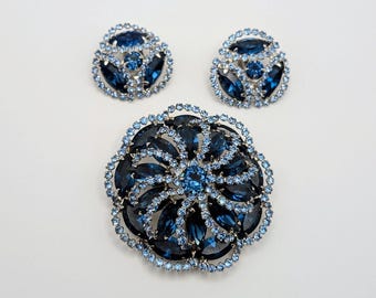 May include: A jewellery set consisting of a circular brooch and two matching earrings. Each piece features dark blue marquise-cut stones arranged in a swirling pattern, surrounded by a border of small, light blue round crystals set in silver.