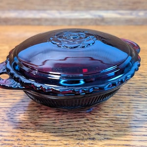 May include: A deep red, covered dish with a floral design on the lid. The dish has a scalloped edge and two handles. The dish is made of glass and sits on a wooden surface.