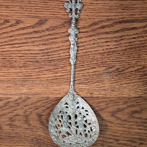 May include: Ornate silver-toned serving spoon with a decorative handle and a pierced bowl. The handle features a fleur-de-lis design at the top and intricate detailing. The bowl has a detailed scene with figures and foliage.