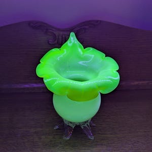 May include: A vibrant, neon green glass vase with a ruffled, flower-like top. The vase has a rounded base and sits on three clear, claw-like feet. The glass glows brightly, creating a striking visual effect.