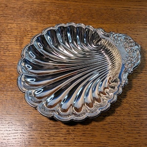 May include: A silver-colored metal dish shaped like a scallop shell. The interior features deep, radiating fluted grooves, while the handle area is decorated with an intricate, embossed floral pattern along the curved edge of the shell.