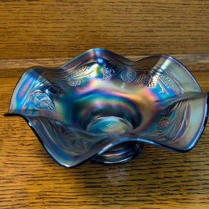 May include: An iridescent blue glass bowl with a ruffled edge. The bowl has a decorative pattern of leaves and wheat. The bowl sits on a wooden surface.