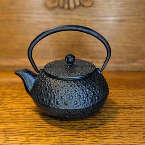 May include: A black cast iron teapot with a textured surface and a curved handle. The teapot has a small spout and a lid with a knob. The teapot is sitting on a wooden surface.