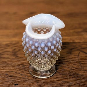 May include: A small, clear glass creamer with a white, ruffled rim and handle. The body of the creamer is textured with small, raised bumps. The creamer sits on a clear, decorative base. The creamer is displayed on a wooden surface.