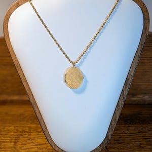 May include: A gold-toned oval locket necklace displayed on a white mannequin. The locket has a textured surface and hangs from a delicate chain. The mannequin is set on a wooden stand, creating a vintage aesthetic.