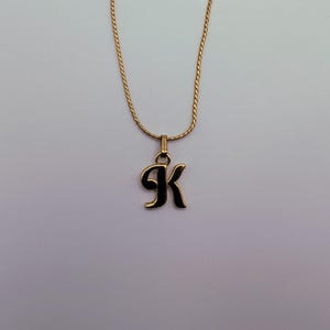 May include: A gold-toned necklace featuring a delicate chain and a pendant shaped like the letter K. The pendant has a smooth, polished finish and is suspended from a simple metal bail, creating a classic piece of jewelry for everyday wear.
