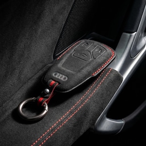 Op de afbeelding: Zwarte autosleutelhoes met rode stiksels en een metalen sleutelhanger. De hoes heeft het Audi-logo en knopiconen. De sleutelhanger ligt op een zwart, suède-achtig oppervlak met rode stiksels.