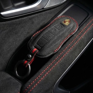 May include: A black leather key fob case with red stitching and a Porsche emblem. The case is resting in a car's interior, next to a door handle and speaker.