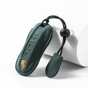 May include: A teal green key fob cover with a black cord and a matching tag. The cover has embossed key symbols and a gold emblem. The fob is made of a soft, suede-like material and is designed to protect the car key.