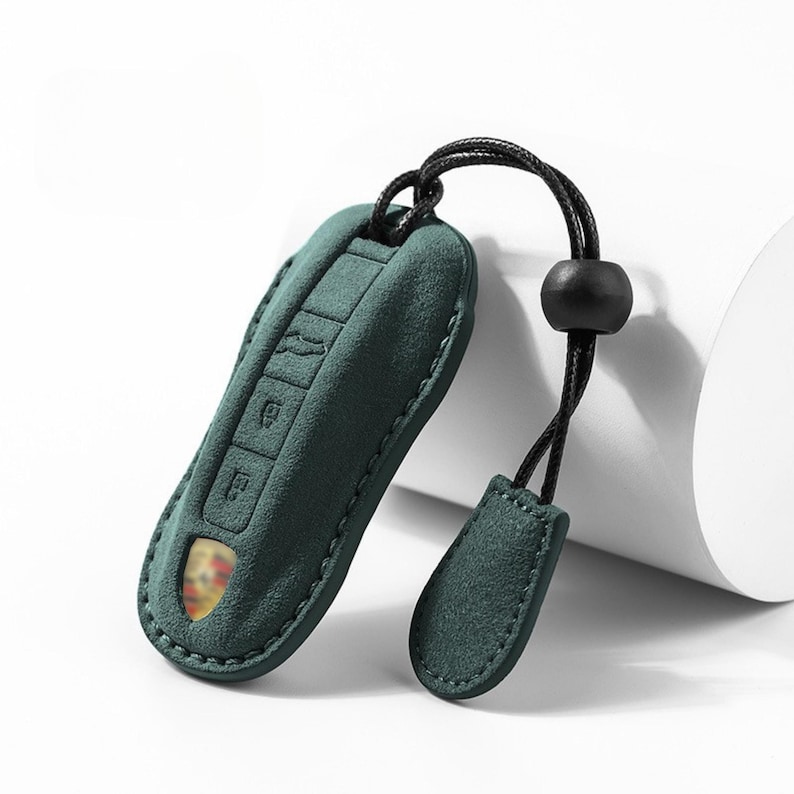 May include: A teal green key fob cover with a black cord and a matching tag. The cover has embossed key symbols and a gold emblem. The fob is made of a soft, suede-like material and is designed to protect the car key.