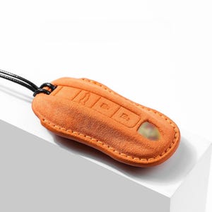 May include: Orange suede car key fob cover with a black cord. The cover has embossed button icons and a keyhole. The stitching is a lighter orange color.