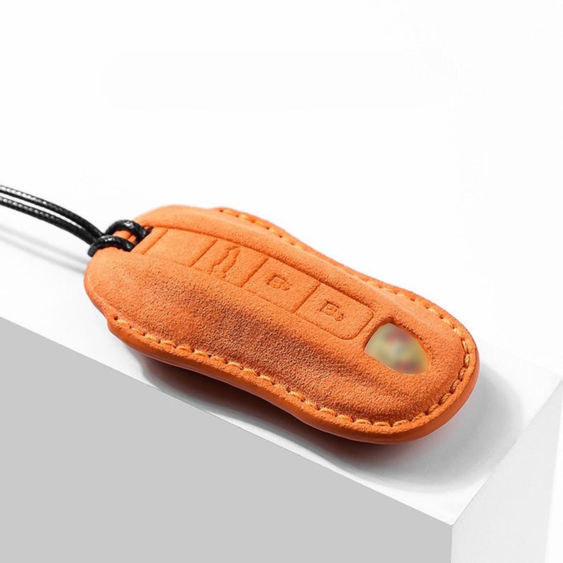 May include: Orange suede car key fob cover with a black cord. The cover has embossed button icons and a keyhole. The stitching is a lighter orange color.
