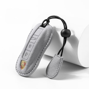 May include: A gray suede key fob cover with a black cord and a black bead. The key fob cover has embossed key symbols and a small emblem. The key fob cover is designed to protect car keys.