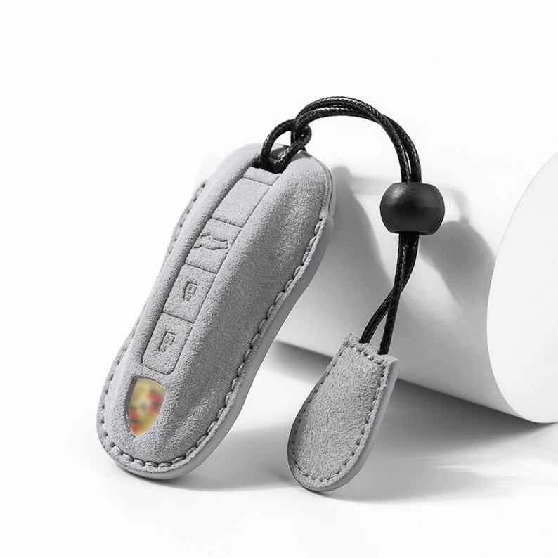 May include: A gray suede key fob cover with a black cord and a black bead. The key fob cover has embossed key symbols and a small emblem. The key fob cover is designed to protect car keys.