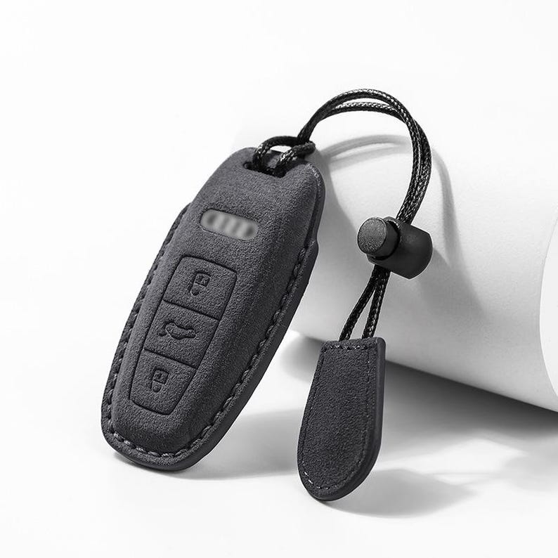 Pode incluir: Uma capa de chave de carro cinza escuro com um cord&atilde;o correspondente e uma pequena etiqueta. A capa da chave tem tr&ecirc;s bot&otilde;es e uma superf&iacute;cie texturizada. O cord&atilde;o tem uma conta preta para ajustar o comprimento.