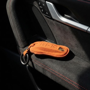 May include: Orange suede car key fob cover with a black metal ring. The key fob cover is resting on a dark gray car interior with red stitching. The key fob cover has embossed key symbols.