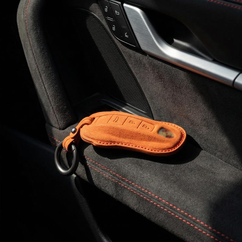 May include: Orange suede car key fob cover with a black metal ring. The key fob cover is resting on a dark gray car interior with red stitching. The key fob cover has embossed key symbols.