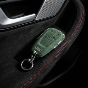 K&ouml;nnte beinhalten: Eine gr&uuml;ne Leder-Autoschl&uuml;sselh&uuml;lle mit wei&szlig;en N&auml;hten und einem silbernen Schl&uuml;sselring. Die H&uuml;lle hat gepr&auml;gte Kn&ouml;pfe und das Audi-Logo. Der Schl&uuml;sselanh&auml;nger liegt in einer schwarzen Autot&uuml;rverkleidung mit roten N&auml;hten.