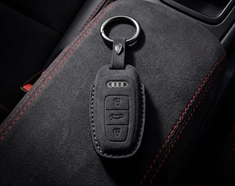 Könnte beinhalten: Ein schwarzer Autoschlüsselanhänger mit Lederetui, mit dem Audi-Logo und den Schlüsseltasten. Der Schlüsselanhänger ist an einem silbernen Schlüsselring befestigt. Der Hintergrund zeigt einen schwarzen Innenraum mit roten Nähten.