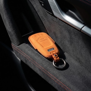 K&ouml;nnte beinhalten: Orangener Wildleder-Autoschl&uuml;sselanh&auml;nger mit silbernem Schl&uuml;sselring. Die Schl&uuml;sselanh&auml;ngerh&uuml;lle hat drei Kn&ouml;pfe und ein Autologo. Die H&uuml;lle liegt in einer schwarzen Autot&uuml;r-Tasche mit roten N&auml;hten.