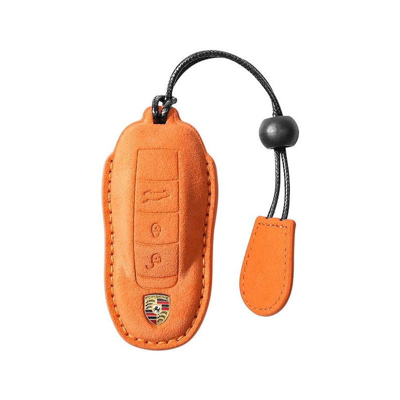 May include: Orange leather key fob cover with a black cord and a small orange tag. The cover has embossed key symbols and a Porsche emblem. The fob is designed to protect a car key.