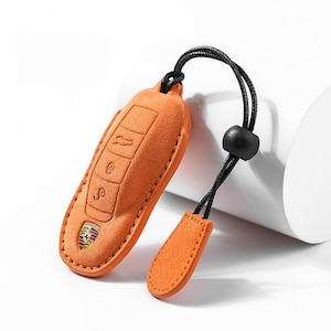 May include: Orange suede key fob cover with embossed key symbols and a Porsche emblem. The cover has a black cord with a black bead and a small orange suede tag.