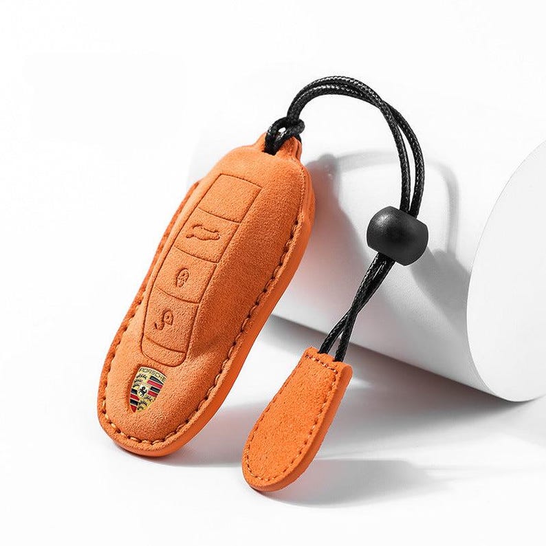 May include: Orange suede key fob cover with embossed key symbols and a Porsche emblem. The cover has a black cord with a black bead and a small orange suede tag.