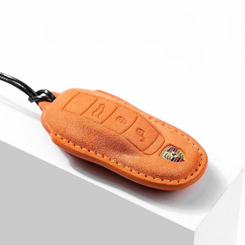 May include: A tan leather car key fob cover with embossed key symbols and a Porsche emblem. The cover is attached to a black cord. The stitching is a contrasting orange color.