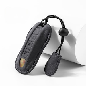 May include: A dark gray key fob cover with a matching lanyard and a small, decorative tag. The cover has embossed key symbols and a stitched border. A gold-colored emblem is visible through a small opening.