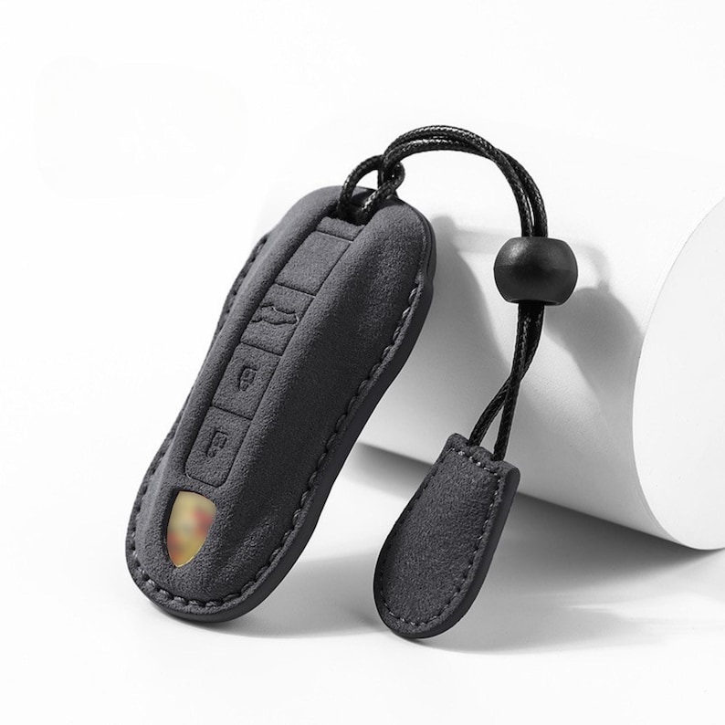 May include: A dark gray key fob cover with a matching lanyard and a small, decorative tag. The cover has embossed key symbols and a stitched border. A gold-colored emblem is visible through a small opening.